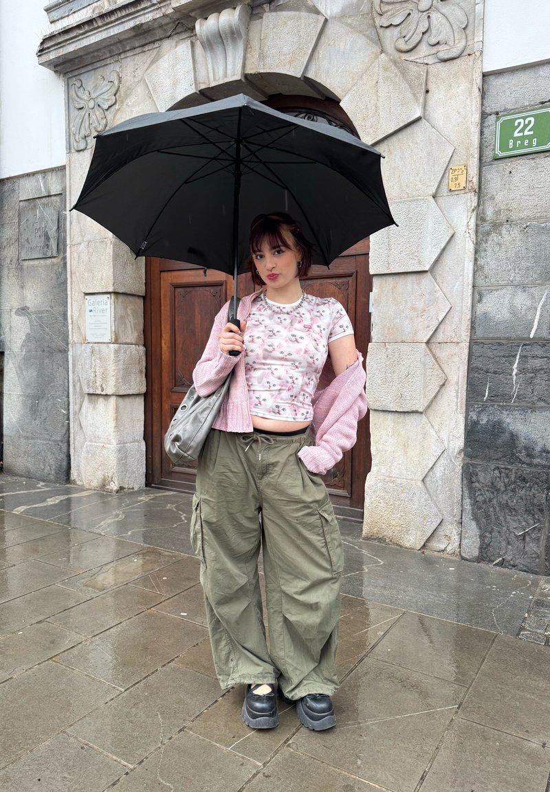 Junge Frau steht auf nassem Pflaster, hält einen schwarzen Regenschirm und trägt eine rosa Strickjacke, ein geblümtes Oberteil, olivgrüne Cargo-Hosen und schwarze Plateau-Schuhe.