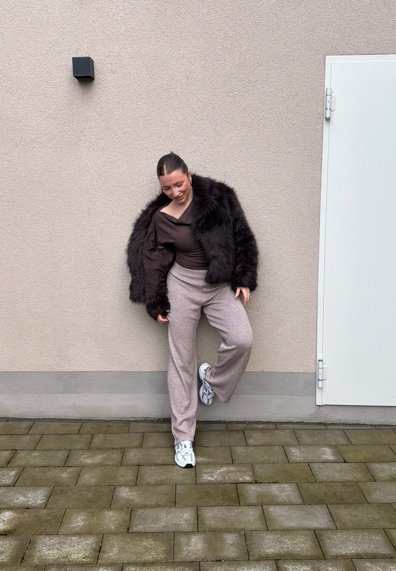Femme en veste en fourrure noire, haut marron, pantalon beige et baskets blanches, appuyée contre un mur gris près d'une porte blanche.