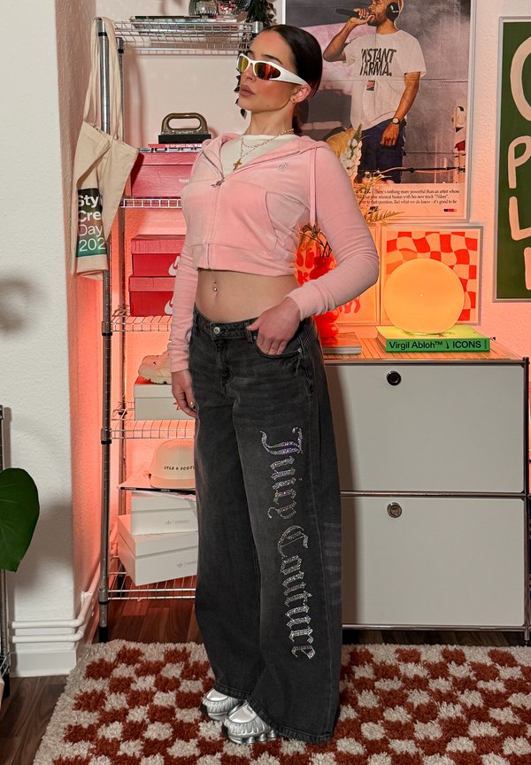 Ženska, ki nosi bele sončna očala, roza skrajšano jopico, črne široke kavbojke z napisom "Juicy Couture", stoji v barviti sobi s policami in plakatom.