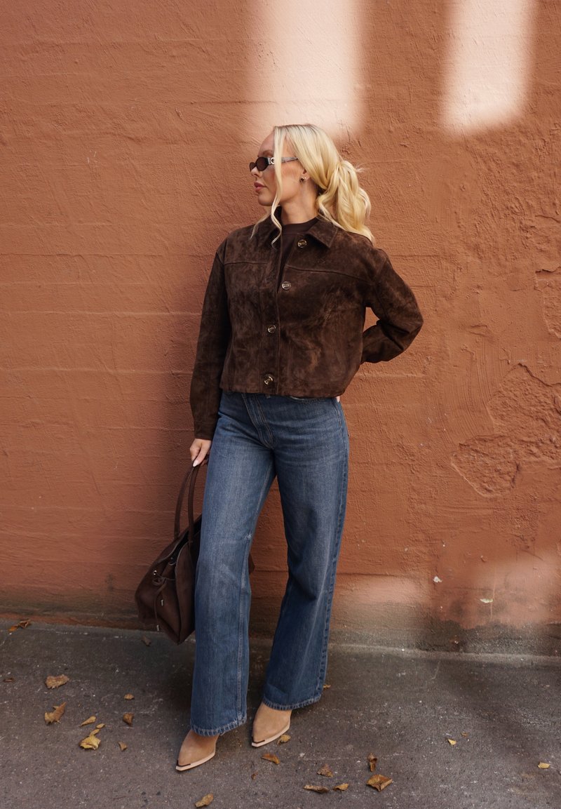 Hnedá semišová krátka bunda so zapínaním na gombíky, kombinovaná so širokými modrými džínsami a béžovými členkovými čižmami s ostrou špičkou, na pozadí textúrovanej steny.