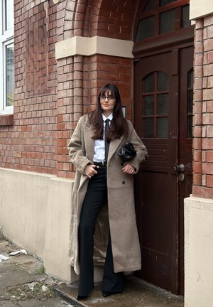 Mujer de cabello largo y oscuro y gafas, vistiendo un abrigo largo beige, camisa blanca, corbata negra y pantalones negros, de pie junto a una pared de ladrillos y una puerta de madera.