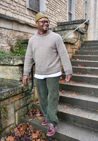 Striped jumper in beige and brown, olive corduroy trousers, maroon shoes, and a green knitted beanie, standing on stone steps with leaves.