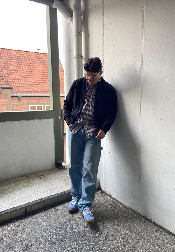 Conjunto casual com um cardigan preto sobre uma camisa às riscas, jeans azul claro e ténis azuis com detalhes brancos, posicionado num canto.