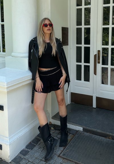 Chaqueta de cuero negra, top negro, falda negra con volantes y botas negras hasta la rodilla. Gafas de sol rojas y detalles plateados con tachuelas en la falda.