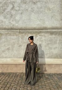 Vrouw in losse, donkerbruine outfit met wijde broek en jas met ceintuur, houdend een kleine olijfgroene handtas, staande op een geplaveide straat.