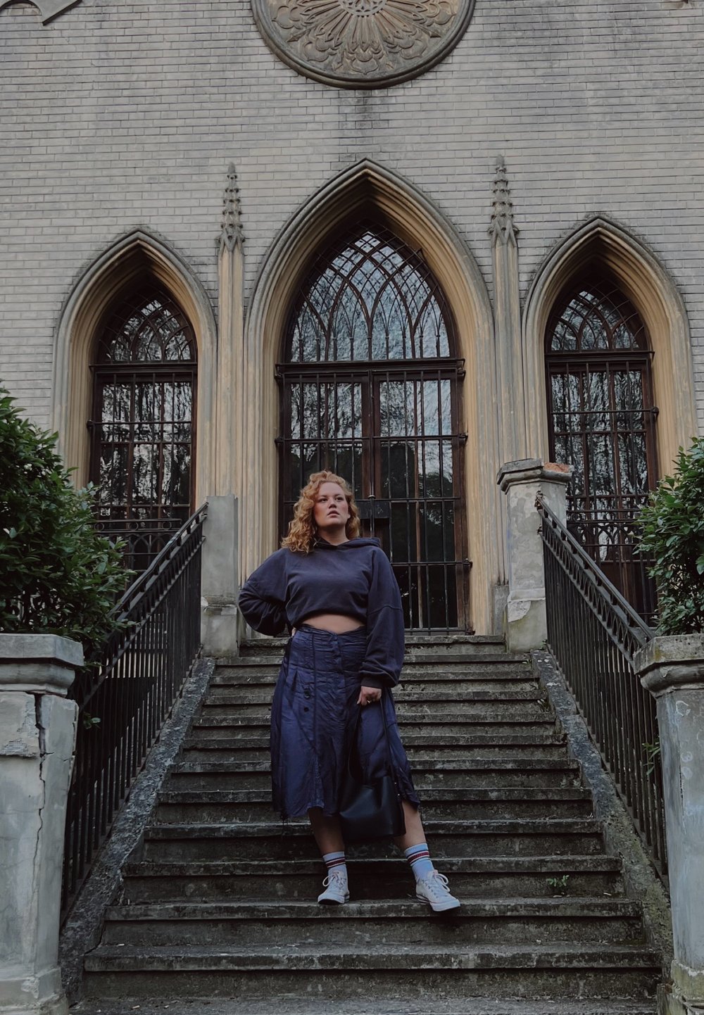 Una donna è in piedi su scale di pietra all'esterno di un edificio di mattoni, indossando un maglione corto blu navy, una lunga gonna e sneaker grigie, mentre tiene una borsa nera.