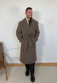 Homme debout à l'intérieur portant un long pardessus marron ceinturé, un pantalon noir et des mocassins en cuir marron foncé avec pompons, souriant avec les mains dans les poches.