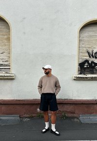 Beige gebreide trui, zwarte op maat gemaakte shorts, witte sokken, zwarte sandalen met witte accenten, staand tegen een gestructureerde lichtgrijze muur.