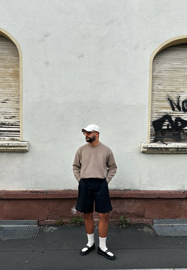 Pullover in beige Strick, schwarze maßgeschneiderte Shorts, weiße Socken, schwarze Sandalen mit weißen Akzenten, stehend vor einer strukturierten hellgrauen Wand.
