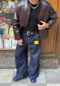 Braune Kunstlederjacke, schwarzes Hemd, weite blaue Jeanshose, silberner Schmuck, gelbes Accessoire am Gürtel befestigt, blaue Schuhe.
