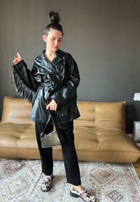 Casaco de cabedal preto com franjas, design de dois botões, com cinto na cintura. Calças pretas e sapatos estampados completam o conjunto. Bolsa minimalista.
