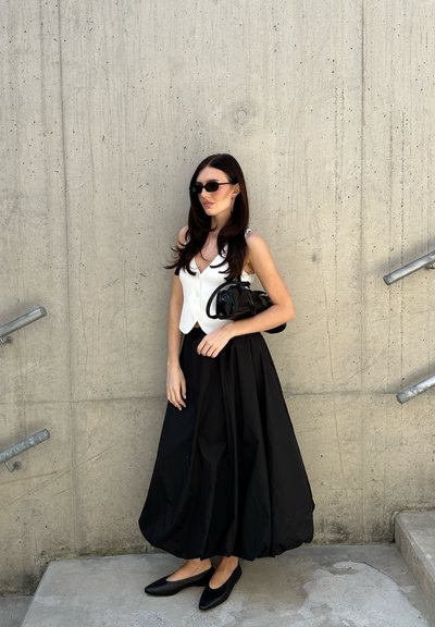 Chaleco blanco ajustado con botones, falda negra vaporosa, zapatos planos negros y un bolso negro. El fondo presenta una pared de concreto texturizada.