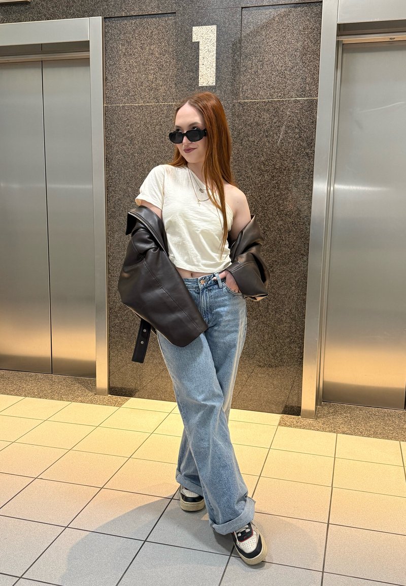 Jeune femme aux longs cheveux roux portant des lunettes de soleil, un t-shirt blanc court, une veste en cuir, un jean large et des baskets, debout entre deux ascenseurs.