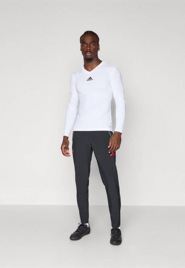 Balta ilgomis rankovėmis sportinė marškinė su juodu Adidas logotipu, derinama su tamsiai pilkomis kelnėmis, turinčiomis raudonus Adidas akcentus, ir juodais sportiniais batais.