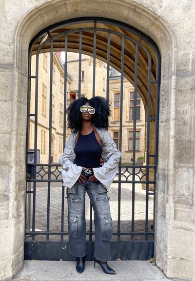 Gafas de sol plateadas de gran tamaño, chaqueta de plumas gris, top negro ajustado y jeans cargo. Botas de tacón negras. Arco de piedra en el fondo.