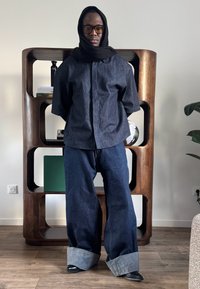 Homme portant une chemise en jean sombre surdimensionnée et un jean large avec revers, avec une écharpe noire sur la tête et des lunettes, debout à l'intérieur près d'une étagère en bois.