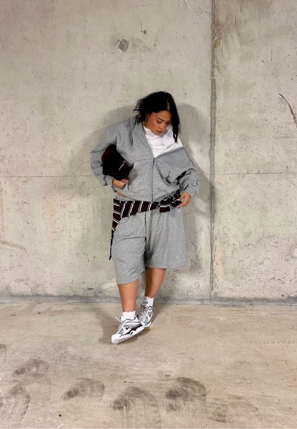 Woman wearing a grey zip-up jacket, matching shorts, white sneakers, and a striped shirt tied around her waist, standing against a concrete wall.