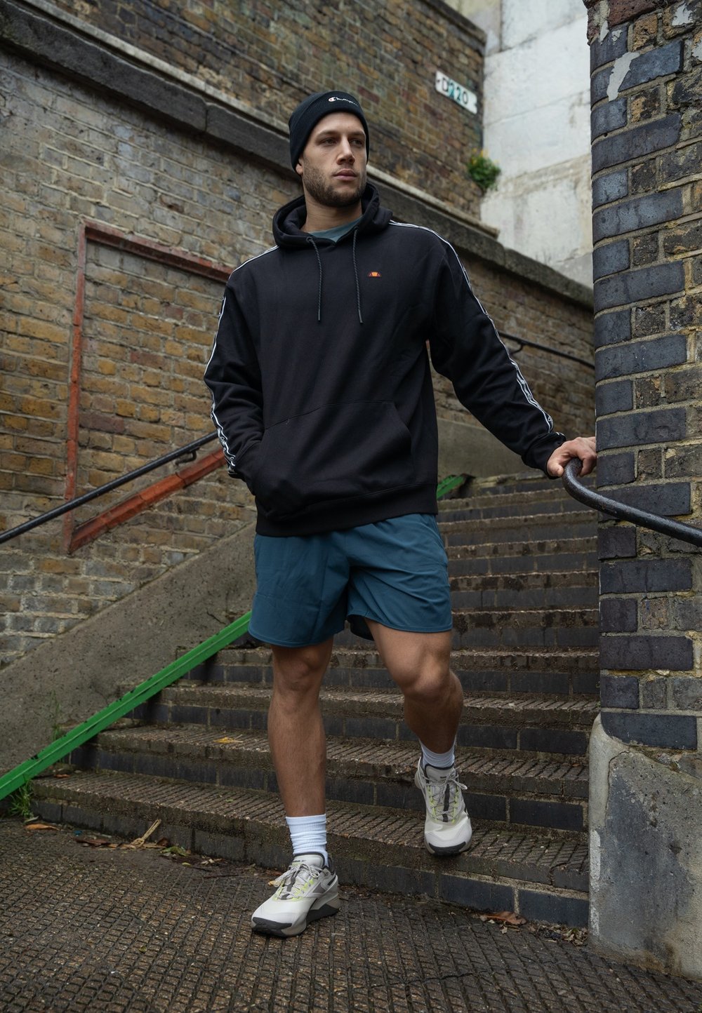 Sudadera con capucha negra y rayas blancas, pantalones cortos color azul petróleo y zapatillas claras. La persona está bajando escaleras de ladrillo con un pasamanos.