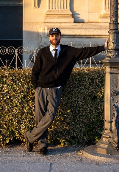 Cardigan negro de canalé sobre una camisa blanca con una corbata negra, pantalones grises y zapatos negros. Lleva una gorra oscura, de pie junto a un farol.
