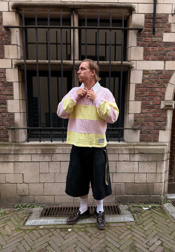 Ung mand med tilbagestrøget hår knapper en pastelrosa og gulstribet langærmet skjorte, stående på en brostensbelagt fortov foran vinduer med gitre.