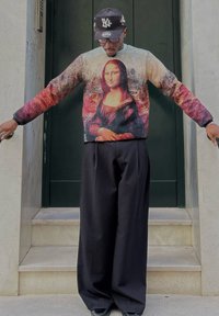 Pull coloré avec l'impression de la Mona Lisa, manches longues, encolure ronde, associé à un pantalon noir ample. Sur fond de porte vert foncé.