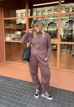 Mujer con chándal mauve de Nike y bolso negro con flecos de pie en una acera con baldosas frente a un edificio con grandes ventanas y paredes naranjas.