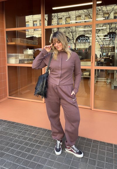 Mujer con chándal mauve de Nike y bolso negro con flecos de pie en una acera con baldosas frente a un edificio con grandes ventanas y paredes naranjas.