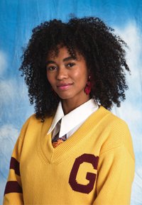 Yellow knitted sweater with a deep V-neck and maroon "G," worn over a white shirt and patterned tie. Curly black hair and red earrings.