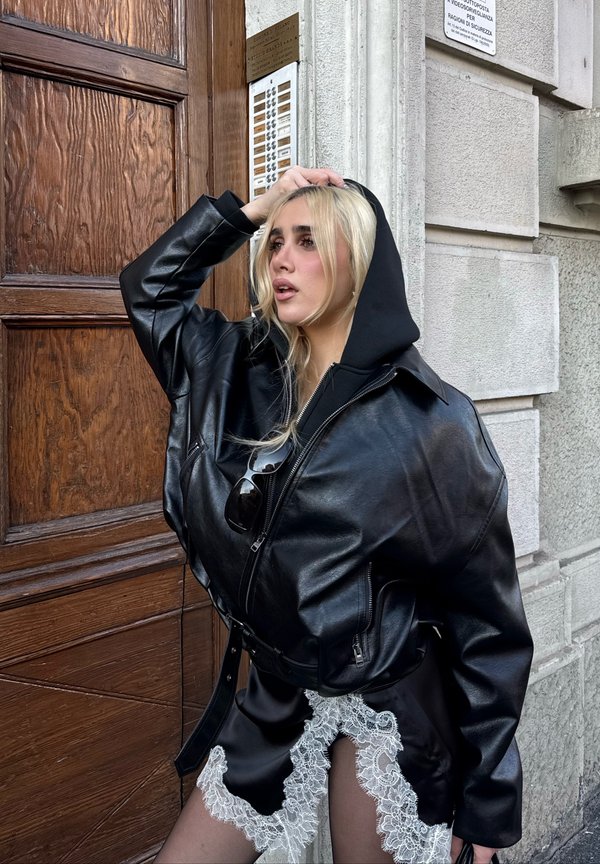 Jeune femme aux cheveux blond platine, vêtue d'une veste en cuir noire et d'une robe ornée de dentelle, posant près d'une porte en bois et d'un mur en pierre à l'extérieur.