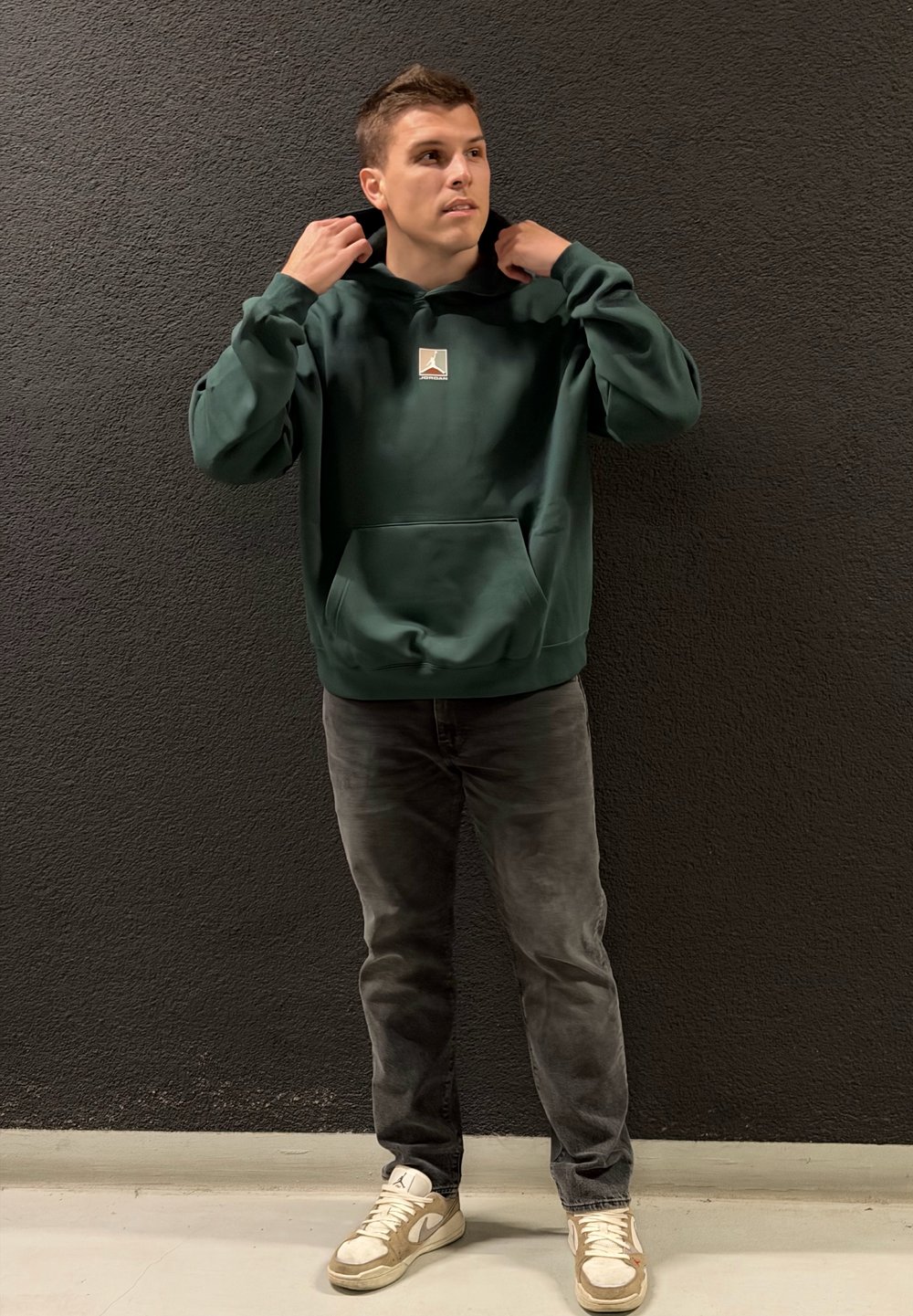 Mørkegrønn hettegenser med kengurulomme og liten logo, kombinert med grå jeans og kremfargede joggesko. Enkelt, uformelt design.