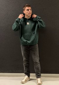 Mørkegrønn hettegenser med kengurulomme og liten logo, kombinert med grå jeans og kremfargede joggesko. Enkelt, uformelt design.