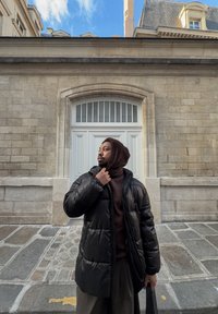 Veste matelassée noire à finition brillante, associée à un sweat à capuche marron texturé. En arrière-plan, un mur en pierre et une porte blanche.