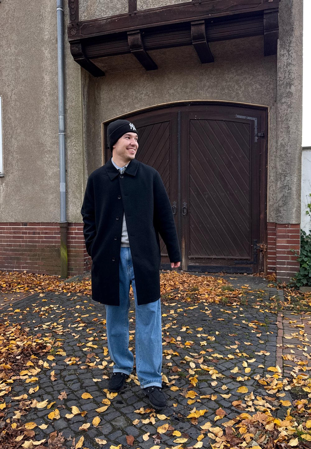 Juodas vilnonis paltas, šviesiai pilkas megztinis, mėlyni džinsai ir juodi sportbačiai. Stovi ant akmeninių grindinių tarp nukritusių geltonų lapų, už nugaros medinės durys.