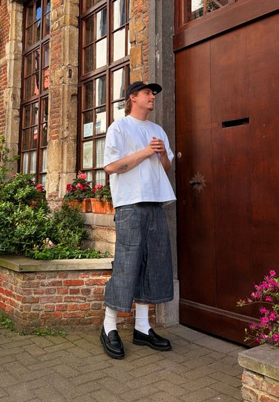 Joven con camiseta blanca, gorra negra y pantalones cortos de mezclilla anchos, de pie con las manos juntas junto a una gran puerta de madera al lado de una pared de ladrillos con plantas.