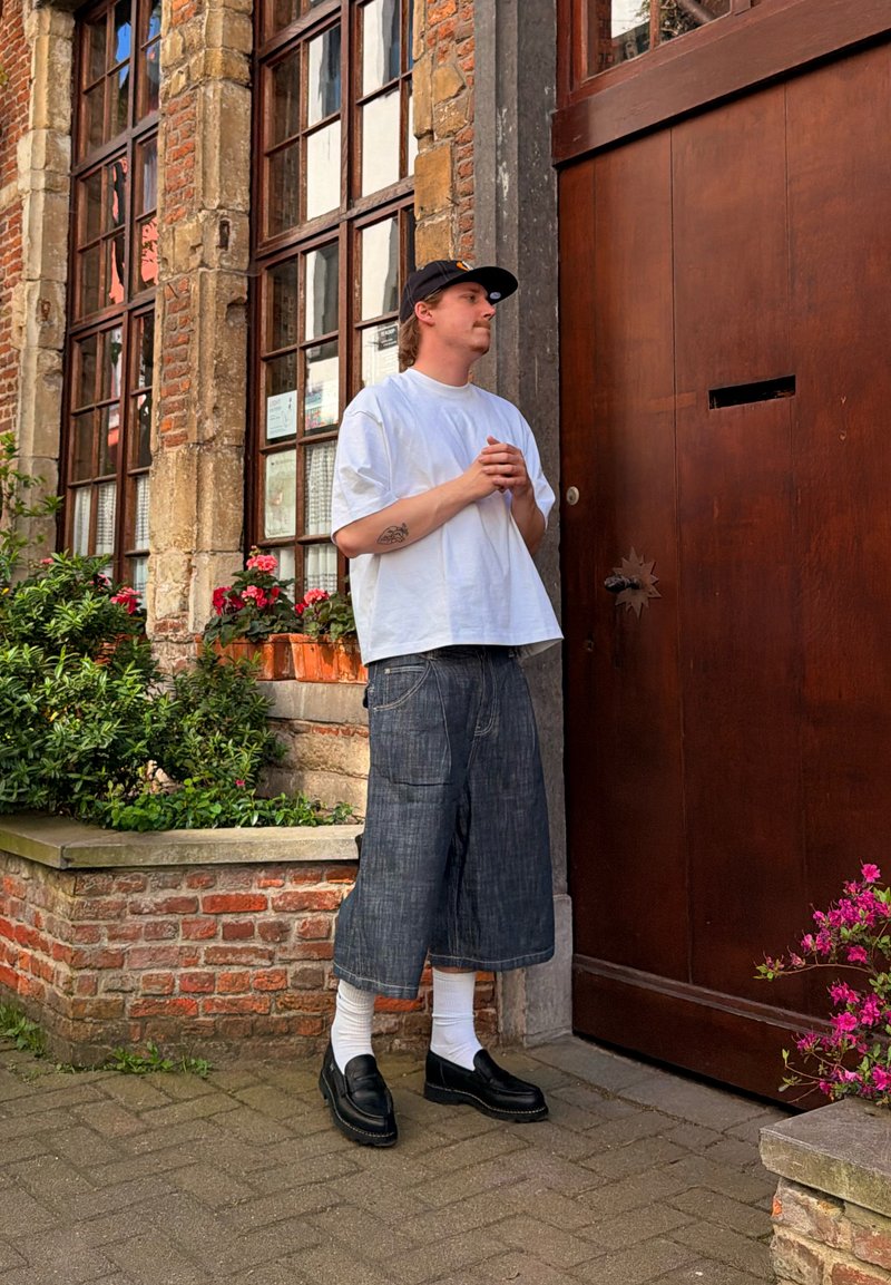 Jeune homme en t-shirt blanc, casquette noire et short en jean large, debout les mains jointes près d'une grande porte en bois à côté d'un mur en briques avec des plantes.