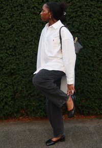 Biała koszula z guzikami z logo, ciemnoszare dżinsy, czarne baleriny i mała czarna torebka na tle zielonego żywopłotu.