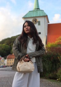 Veste kaki sur un haut en tricot blanc, associée à un pantalon blanc ample. Tient un sac à main beige texturé avec des poignées marron.
