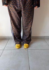 Satin hlače z leopardjim vzorcem in črnimi ter belimi stranskimi črtami, v kombinaciji z svetlo rumenimi čevlji s vezalkami. Postavljene na ploščicami obloženem tleh.