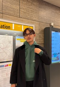 Homme en sweat-shirt vert sous un manteau noir, debout près d'une carte des transports et d'un tableau des horaires dans une station.
