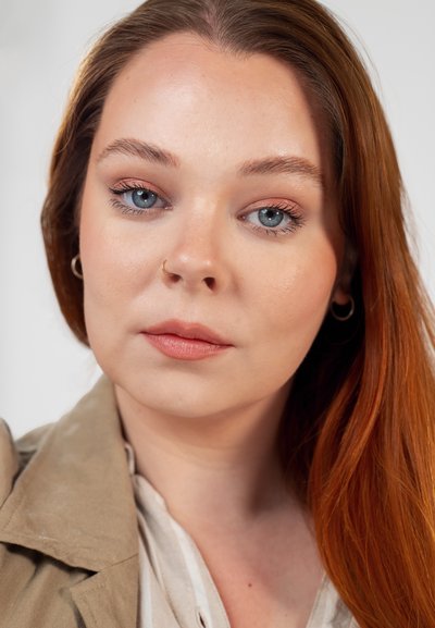 Joven mujer con cabello largo y rojo, maquillaje ligero, anillo en la nariz y pendientes, vistiendo una chaqueta beige y blusa blanca sobre un fondo liso.