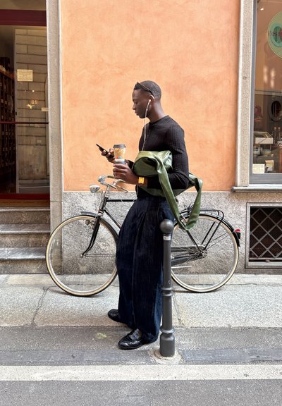 Persona vestida con un suéter negro de canalé y pantalones anchos oscuros. Sosteniendo una taza de café y una bolsa verde, de pie junto a una bicicleta negra.