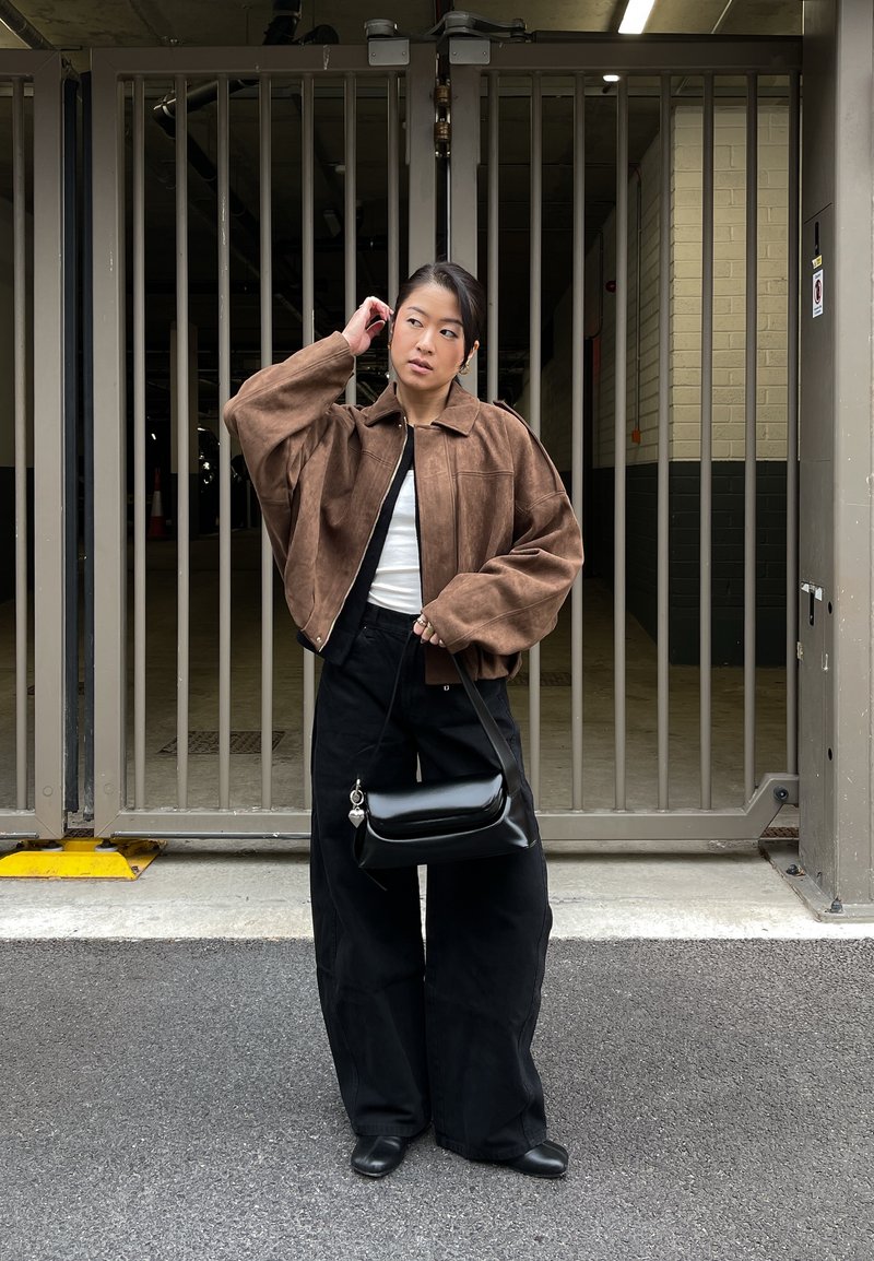 Veste courte en daim marron, haut blanc, pantalon large noir, sac à épaule noir avec détails métalliques, debout devant une zone clôturée.