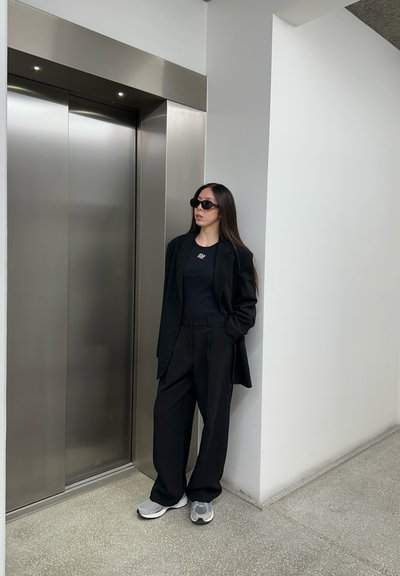 Joven mujer con traje negro y zapatillas, usando gafas de sol, de pie cerca de puertas cerradas de ascensor de acero inoxidable en un pasillo interior moderno.