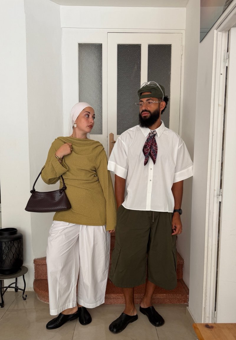 Deux personnes debout à l'intérieur près de portes blanches, l'une portant un hijab, un pull vert, un pantalon blanc, l'autre avec une casquette, une chemise blanche, un short vert, tous deux avec des chaussures noires.
