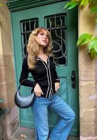 Cardigan nero a coste con piping bianco, abbottonato, abbinato a jeans a zampa d'elefante blu chiaro; una borsa a forma di mezzaluna nera è tenuta in mano. Sfondo di una porta verde.