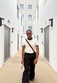 Hvit T-skjorte med svart kant, svarte vide bukser og brun skulderveske. Utendørs korridor med lyse vegger og store vinduer.