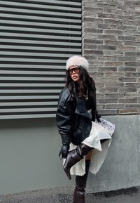 Schwarze übergroße Lederjacke, weißes Kleid mit Spitzenbesatz, kniehohe braune Stiefel, flauschiger weißer Hut, Sonnenbrille, braune Handschuhe, strukturierter Hintergrund.