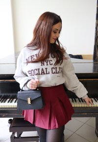 Felpa grigia con il testo "NEW YORK NYC SOCIAL CLUB", gonna plissettata bordeaux, borsa nera con hardware dorato e collant neri.