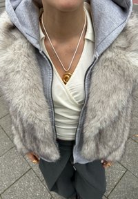 Grå faux-fur vest med hette, brukt over en kremfarget topp. En gyllen spiralanheng henger fra en hvit snor. Mørkegrå bukser er synlige.