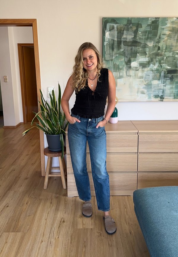 Femme aux cheveux blonds bouclés portant un haut noir sans manches, un jean bleu et des pantoufles, debout dans le salon avec une plante et une peinture abstraite.